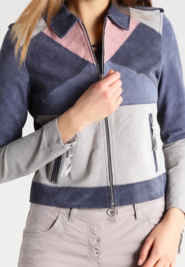Femme portant une veste en suède color-block bleu marine, gris et rose, avec une fermeture éclair devant et des poches zippées, ajustant la tirette de la fermeture éclair.