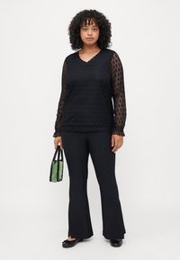 Μαύρη δαντέλα με μακρύ μανίκι και υφή με σχέδιο, συνδυασμένη με μαύρα φαρδιά παντελόνια και παπούτσια. Κρατώντας μια τσάντα από χάντρες σε πράσινο και μαύρο χρώμα.