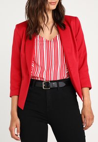 Blazer rosso con revers, abbinato a una blusa a righe rosse e bianche. Una cintura nera e pantaloni neri aderenti completano l'outfit.