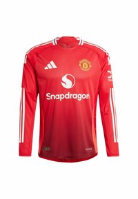 Jersey rouge à manches longues en matériau léger, avec des rayures blanches sur les épaules, un col rond et le logo de Manchester United sur la poitrine.