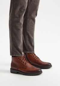 Brązowe skórzane botki za kostkę z okrągłymi noskami, sznurowanym zapięciem i teksturowanymi gumowymi podeszwami, zestawione z szarymi spodniami z kordurowej tkaniny.