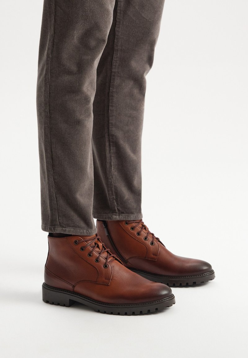 Brązowe skórzane botki za kostkę z okrągłymi noskami, sznurowanym zapięciem i teksturowanymi gumowymi podeszwami, zestawione z szarymi spodniami z kordurowej tkaniny.