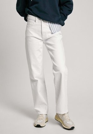 Person trägt gerade geschnittene weiße Jeans, beige und weiße Sneakers, einen marineblauen Pullover und ein gestreiftes Hemd, das teilweise herausgezogen ist.