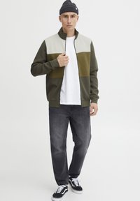 Giacca verde e grigia con zip e colletto alto, caratterizzata da blocchi di colore. Abbinata a jeans scuri e sneakers nere. Design semplice.