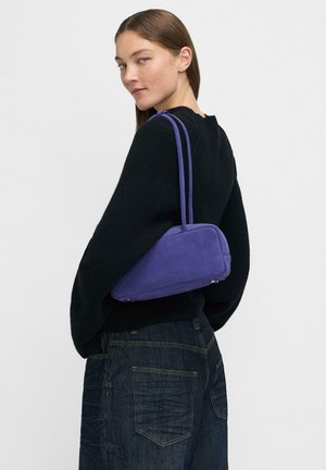 Borsa a tracolla in suede viola con design strutturato, dotata di due cinturini sottili e dettagli in metallo argentato. Indossata sopra un maglione nero.