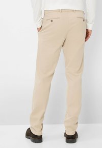Chino beige con vestibilità dritta, dotati di tasche posteriori con chiusura a bottone e una texture di tessuto liscia. Indossati con scarpe marrone scuro.