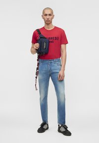Άνδρας που φοράει κόκκινο γραφικό T-shirt, ανοιχτό μπλε τζιν, μαύρα αθλητικά παπούτσια και κρατά μια ναυτικού μπλε τσάντα crossbody με λουρί στον ώμο.