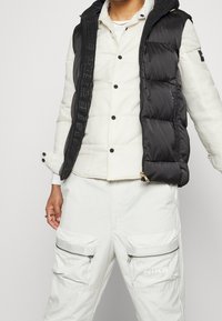 Osoba ubrana w białą pikowaną kurtkę i białe spodnie cargo Nike, z czarną kurtką puchową na wierzchu.