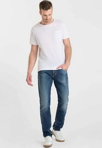 Witte T-shirt met ronde hals en zakje, gecombineerd met blauwe spijkerbroek. Model draagt witte sneakers. Eenvoudig, casual ontwerp met een ontspannen pasvorm.