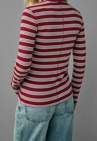 Femme portant un haut à manches longues ajusté à rayures bordeaux, blanches et bleues, avec un jean en denim bleu clair, debout devant un fond gris.