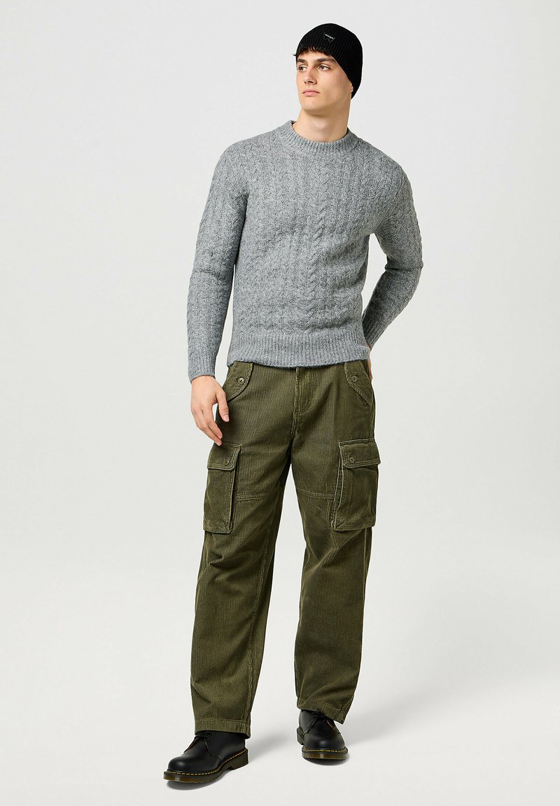 Suéter de punto gris, pantalones cargo de pana verde oliva con bolsillos, y botas negras con cordones. El atuendo tiene una apariencia casual y texturizada.