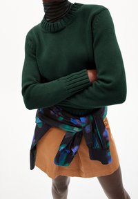Pull en tricot vert avec un col côtelé, associé à une jupe marron et une écharpe noire ornée de motifs verts et bleus.