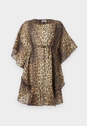 Mladá žena stojí s rukou na boku, oblečená v priesvitných šatách so vzorom leopard a čiernych sandáloch na podpätku na bielom pozadí.