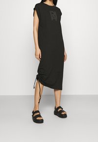 Czarna sukienka do kolan z krótkimi rękawami, z logiem na piersi i bocznymi sznurkami. Połączona z płaskimi czarnymi sandałami.