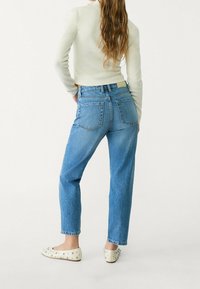 Mujer con cabello largo y ondulado vestida con un suéter claro, jeans azul de cintura alta y bailarinas blancas con tachuelas, de pie con las manos en las caderas mirando hacia atrás.