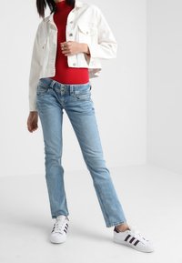Bílá džínová bunda, červený fitness top, modré slim džíny a bílé tenisky s fialovými pruhy. Neformální outfit na jednobarevném pozadí.
