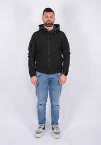 Giacca nera con zip e cappuccio verde, abbinata a jeans azzurri chiari e scarpe da ginnastica bianche. Presenta un design elegante e tasche laterali.