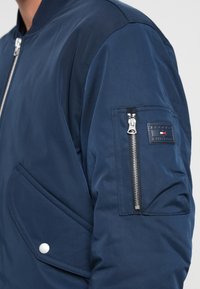 Navy blue bomber jacket with front snap pocket, silver zipper on sleeve pocket, and brand logo patch on upper arm.