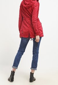 Rood gevoerde parka met verstelbare trekkoord taille, textuur zijpanelen en voorzakken, gecombineerd met omgeslagen blauwe spijkerbroek en zwarte enkelboots.