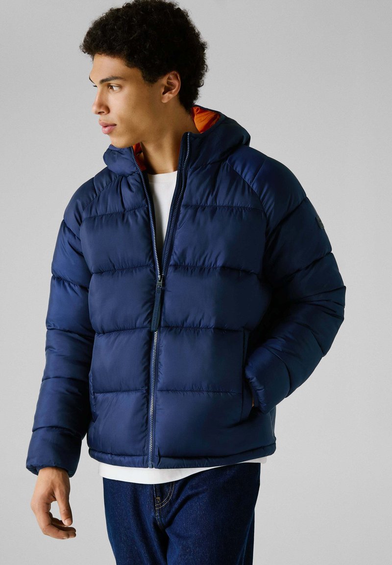 Marineblauwe pufferjack met een capuchon, met een gecapitonneerd ontwerp, een rits aan de voorkant en oranje voering. Het materiaal lijkt glad met een glanzende afwerking.