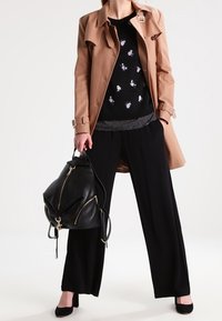 Trench-coat beige porté sur un pull noir à motifs floraux, associé à un pantalon noir à jambes larges et un sac à dos noir texturé. Des chaussures noires complètent la tenue.