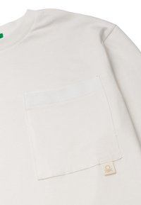 Bílý bavlněný tričko s kapsou na prsou a malou značkovou nálepkou. Tkanina má hladkou texturu s klasickým designem kulatého výstřihu.