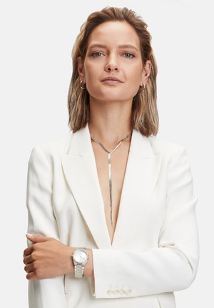 Silberner Anhänger-Halsreifen mit einer schlanken, länglichen Form, getragen über einem tief ausgeschnittenen weißen Blazer. Silberne Uhr mit Metallarmband am Handgelenk.