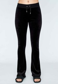 Zwarte fluwelen flared broek met een elastische tailleband en trekkoord. Heeft een gladde textuur en subtiele glans, gecombineerd met casual sandalen.