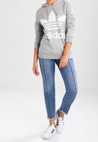 Grå hoodie med vitt Adidas-logotyp, blå jeans med fransad nederkant och vita sneakers med svarta ränder. Mjukt bomullsmaterial, avslappnad passform.