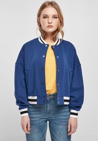 Blue bomber jacket with a button-up front, white and black striped trim on cuffs and hem, worn over a yellow shirt and blue jeans.