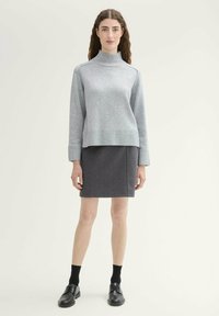 Pull en col roulé gris clair avec un ourlet et des poignets côtelés, associé à une jupe à motif écossais sombre. Des chaussures noires complètent la tenue.
