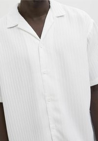 Persona di carnagione scura che indossa una camicia bianca a maniche corte con bottoni, caratterizzata da un motivo a righe verticali texturizzate e un colletto alla carlona.