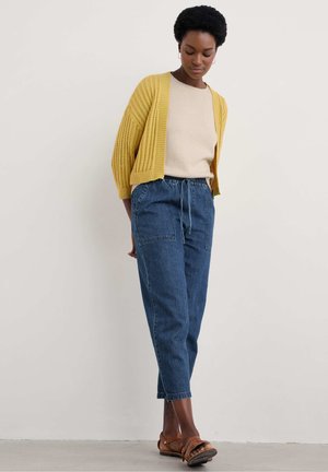 Cardigan giallo lavorato a maglia sopra un maglione beige, abbinato a pantaloni di jeans blu larghi e sandali marroni. Tessuto testurizzato, vestibilità rilassata.