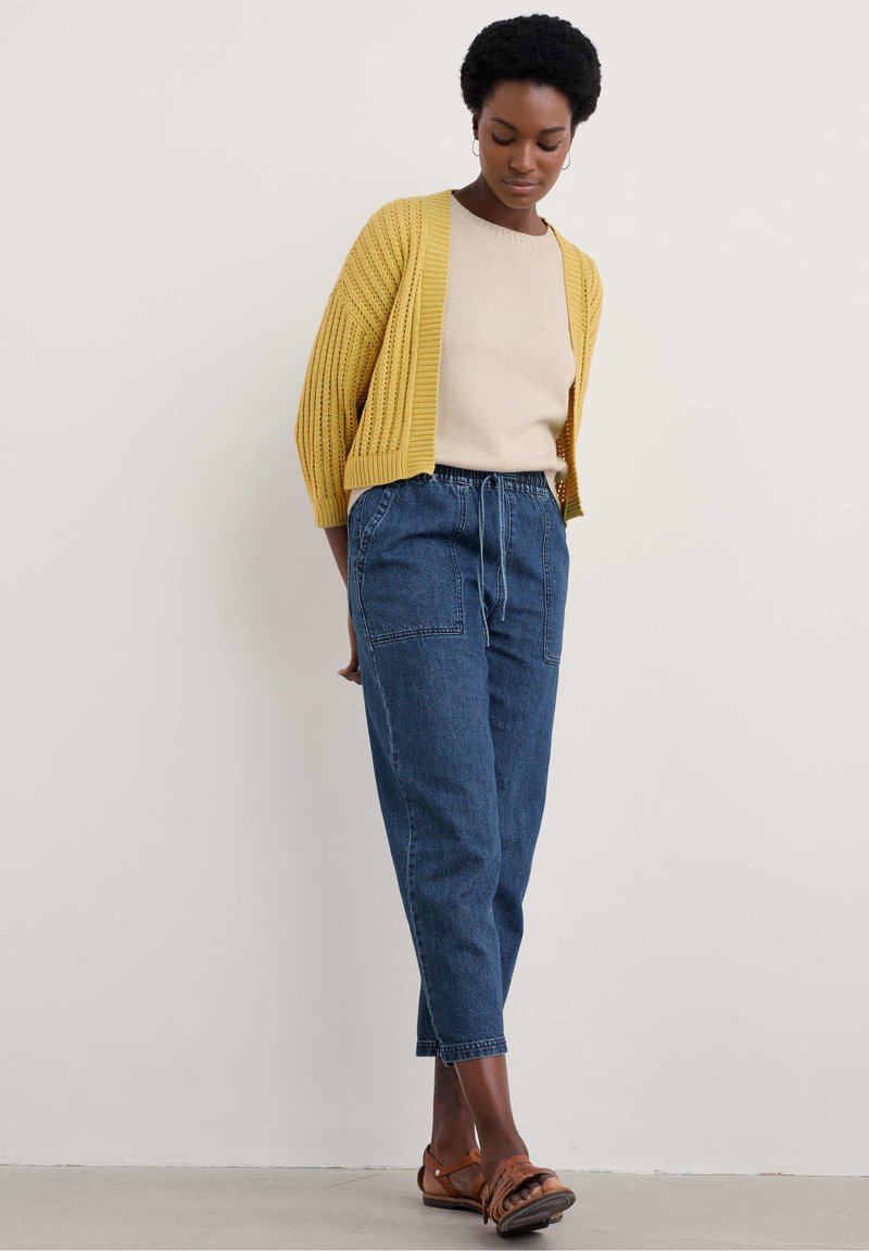 Gelber Strickcardigan über beigem Pullover, kombiniert mit lockeren blauen Denim-Hosen und braunen Sandalen. Texturiert, lockere Passform.
