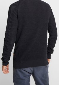 Man som bär en mörkgrå ribbstickad tröja med strukturerade ärmar och blågrå byxor, sedd bakifrån.