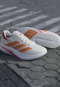 Baskets blanches avec des bandes orange et des accents rouges au talon posées sur un pavé en pierre texturé à proximité d'un passage piéton.