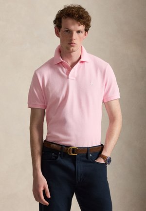 Giovane uomo con capelli ricci che indossa una polo rosa chiaro, pantaloni scuri, cintura marrone e orologio da polso, in piedi davanti a uno sfondo semplice.