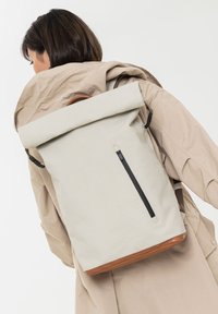 Beige rugzak met oprolbare bovenkant, gemaakt van gladde stof, zwarte ritssluiting en een bruine leren onderkant. Voorzien van verstelbare riemen voor een goede pasvorm.