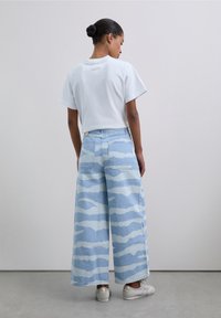 White cropped t-shirt, loose-fitting light blue wide-leg jeans with wavy light blue pattern. Model wears silver sneakers. Plain background.