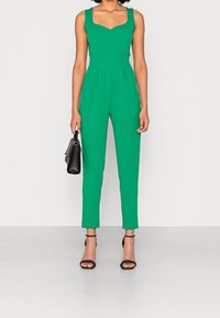 Green sleeveless jumpsuit with a sweetheart neckline, fitted bodice, pleats, and tapered leg; paired with black strappy heels and a handbag.