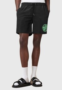 Bañador negro con cinturilla elástica y cordón, con un detalle gráfico circular verde. Lleva sandalias negras y calcetines blancos.