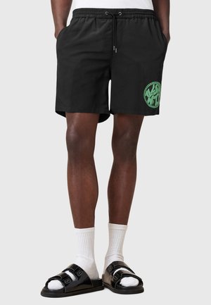 Pantaloni da nuoto neri con vita elasticizzata e cordino, caratterizzati da un dettaglio grafico circolare verde. Indossati con sandali neri e calzini bianchi.