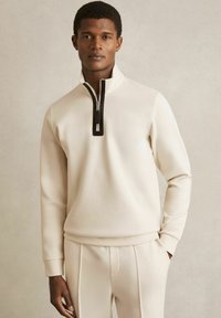 Sweatshirt de culoare crem, cu guler înalt și închidere cu fermoar negru. Fabricați dintr-un material moale, având mâneci lungi și o croială lejeră.
