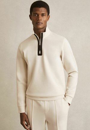 Sweatshirt de culoare crem, cu guler înalt și închidere cu fermoar negru. Fabricați dintr-un material moale, având mâneci lungi și o croială lejeră.