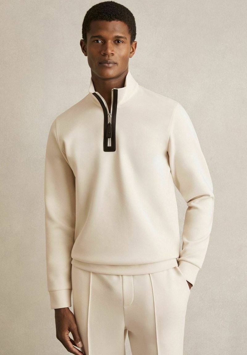 Sweatshirt de culoare crem, cu guler înalt și închidere cu fermoar negru. Fabricați dintr-un material moale, având mâneci lungi și o croială lejeră.