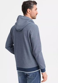 Sweat-shirt à capuche en tissu gris foncé avec une texture lisse, présentant une bordure et des poignets côtes de couleur marine. Le modèle montre la vue arrière.