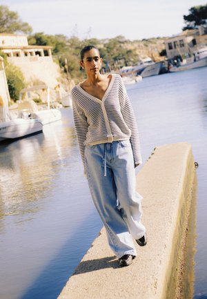Vrouw in een gestreepte cardigan en wijde spijkerbroek die langs een smalle betonnen steiger bij een jachthaven met boten en gebouwen op de achtergrond loopt.