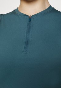 Blågrøn sportstop i glat, elastisk materiale med kort lynlås ved halsen og rundet halsudskæring. Ingen mønstre eller dekorationer synlige.