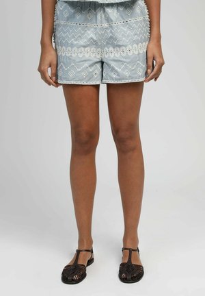 Personne portant un short bleu clair avec des broderies géométriques et florales blanches, et des sandales tressées à bout fermé marron foncé.