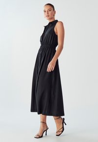 Juoda be rankovių midi suknelė su mygtukų užsegimu priekyje, susirinkusiu elastingu juosmeniu ir laisvai krentančia sijonu. Su juodais užsegamais aukštakulniais.